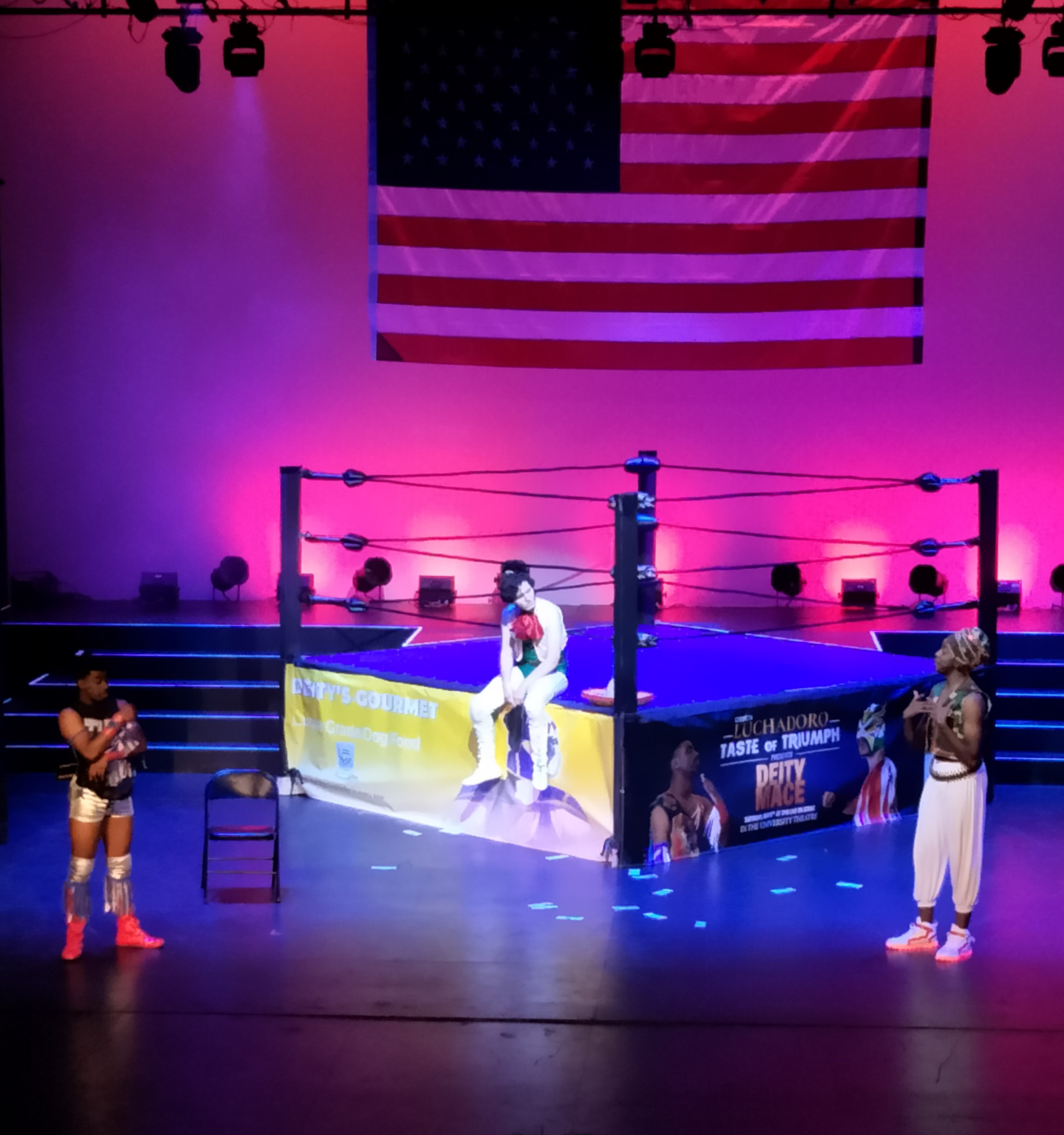 Photo of actors around a wrestling ring bathed in pink and purple light.