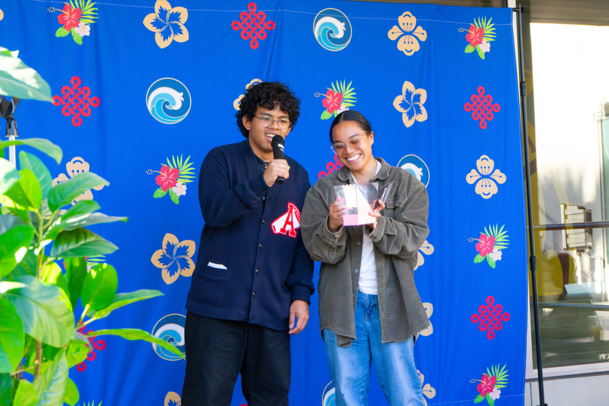 Two students smile on stage during an event, one speaking into a mic while the other holds a raffle box in front of a blue floral backdrop