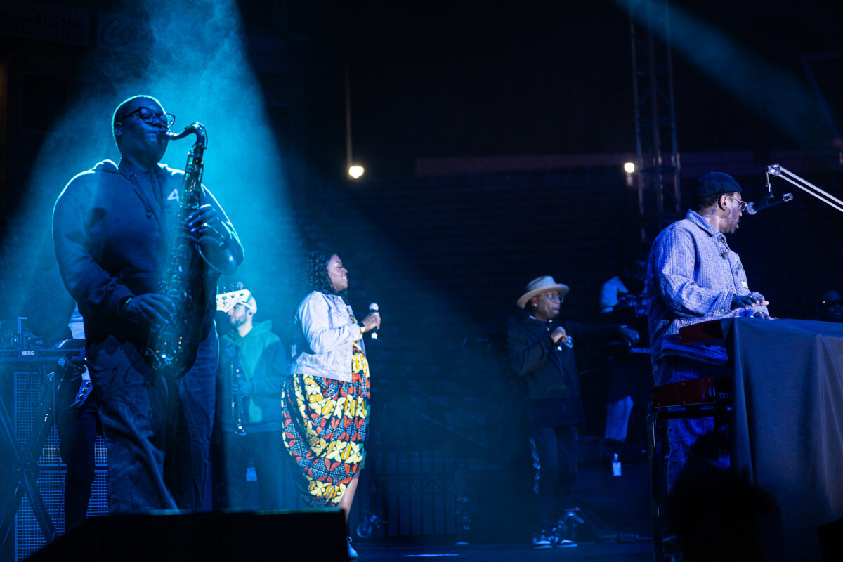 Musicians perform on stage under blue lighting, with a saxophonist in the spotlight and vocalists and a keyboard player in the background.