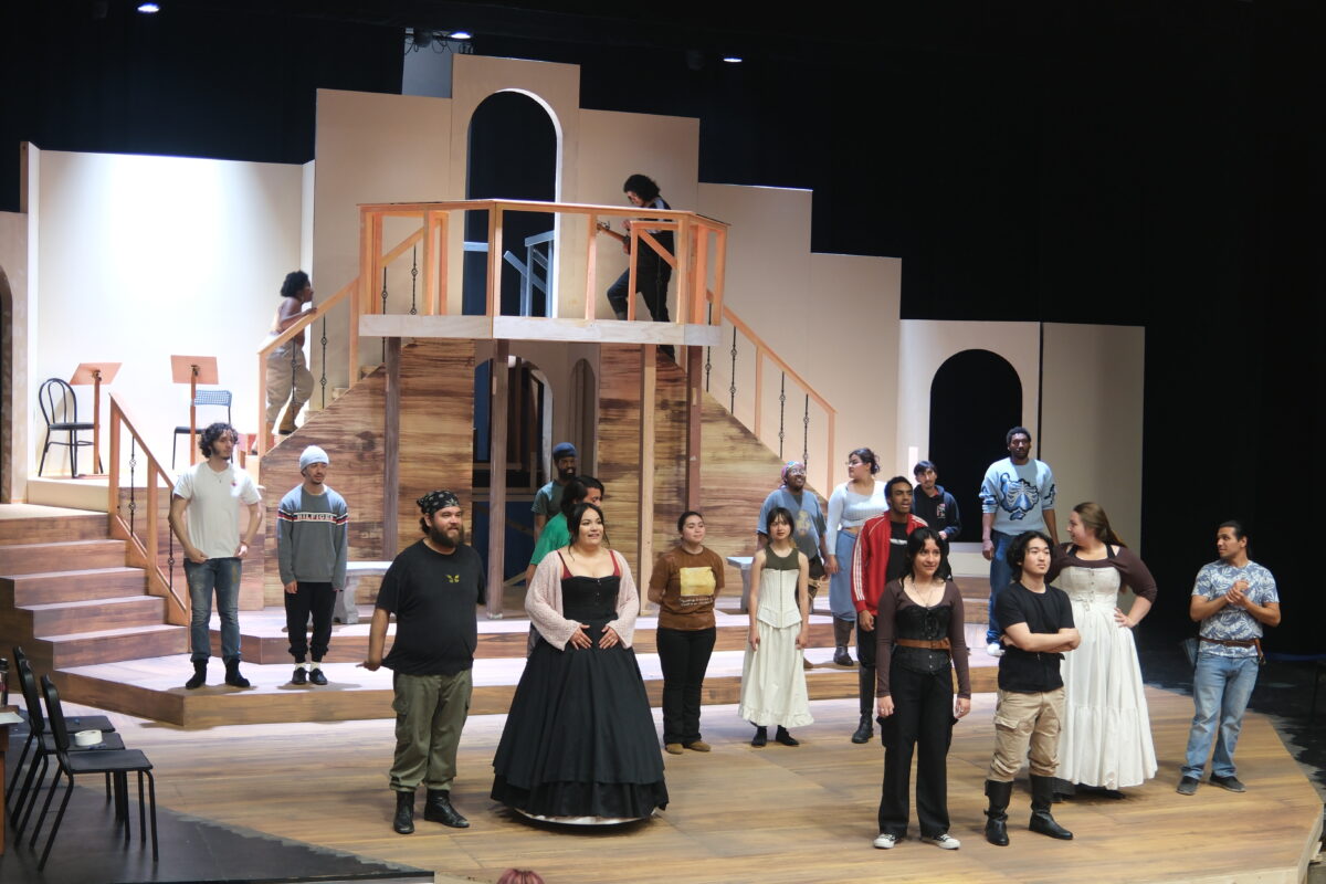 Theater students rehearse on a wooden stage set with stairs and balconies, wearing costumes and standing in various positions across the stage.