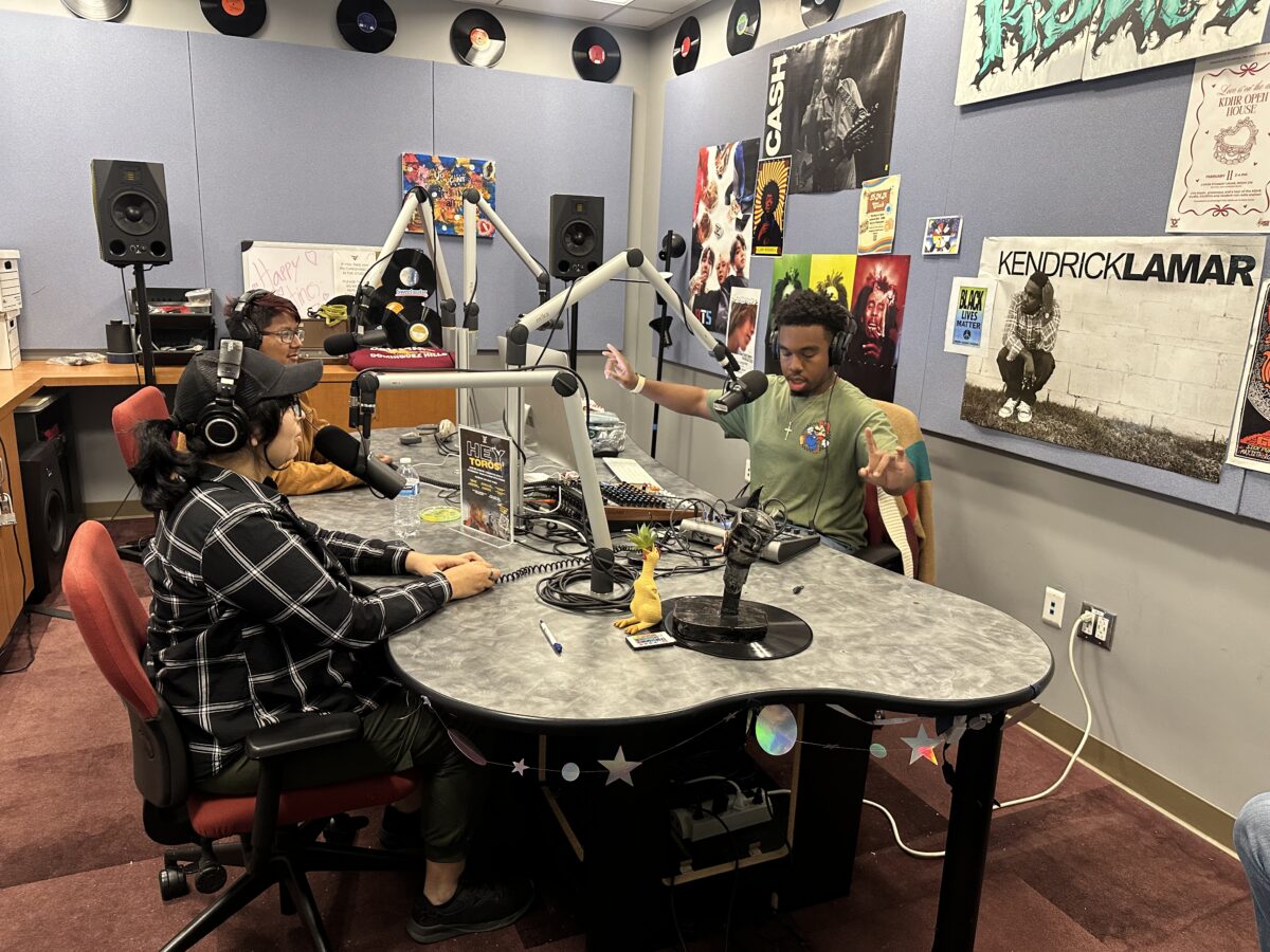 Three students sit in a radio studio with microphones during a recording session of a student-run game show.
