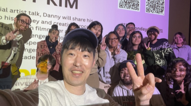 Danny Kim takes a selfie with a group of students in front of a projected screen during his guest artist talk, with several people making peace signs and smiling.