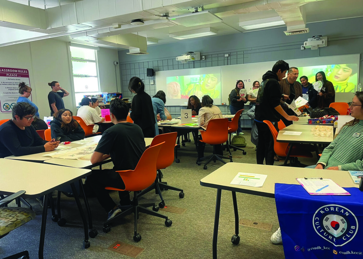 Students gather in a classroom for a Korean Culture Club event, seated at tables doing activities while K-pop videos play on the projector screen.