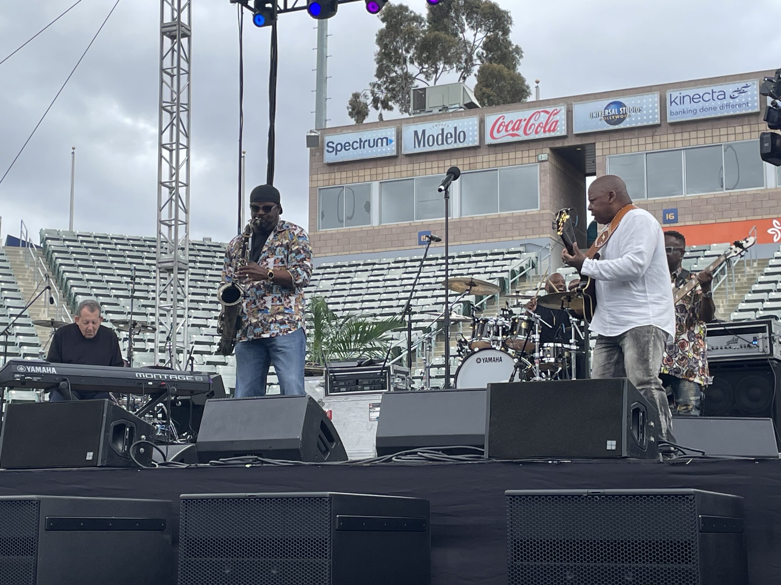 A jazz band performs on an outdoor stage during the day, with musicians playing keyboard, saxophone, guitar, bass, and drums in front of empty stadium seats.
