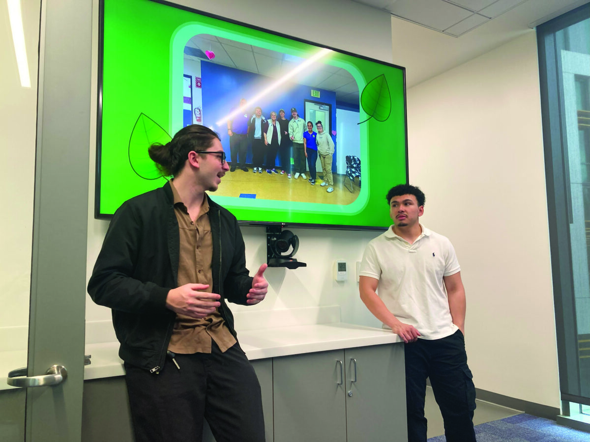 Two students present in a classroom, standing in front of a screen showing a group photo with a green border.