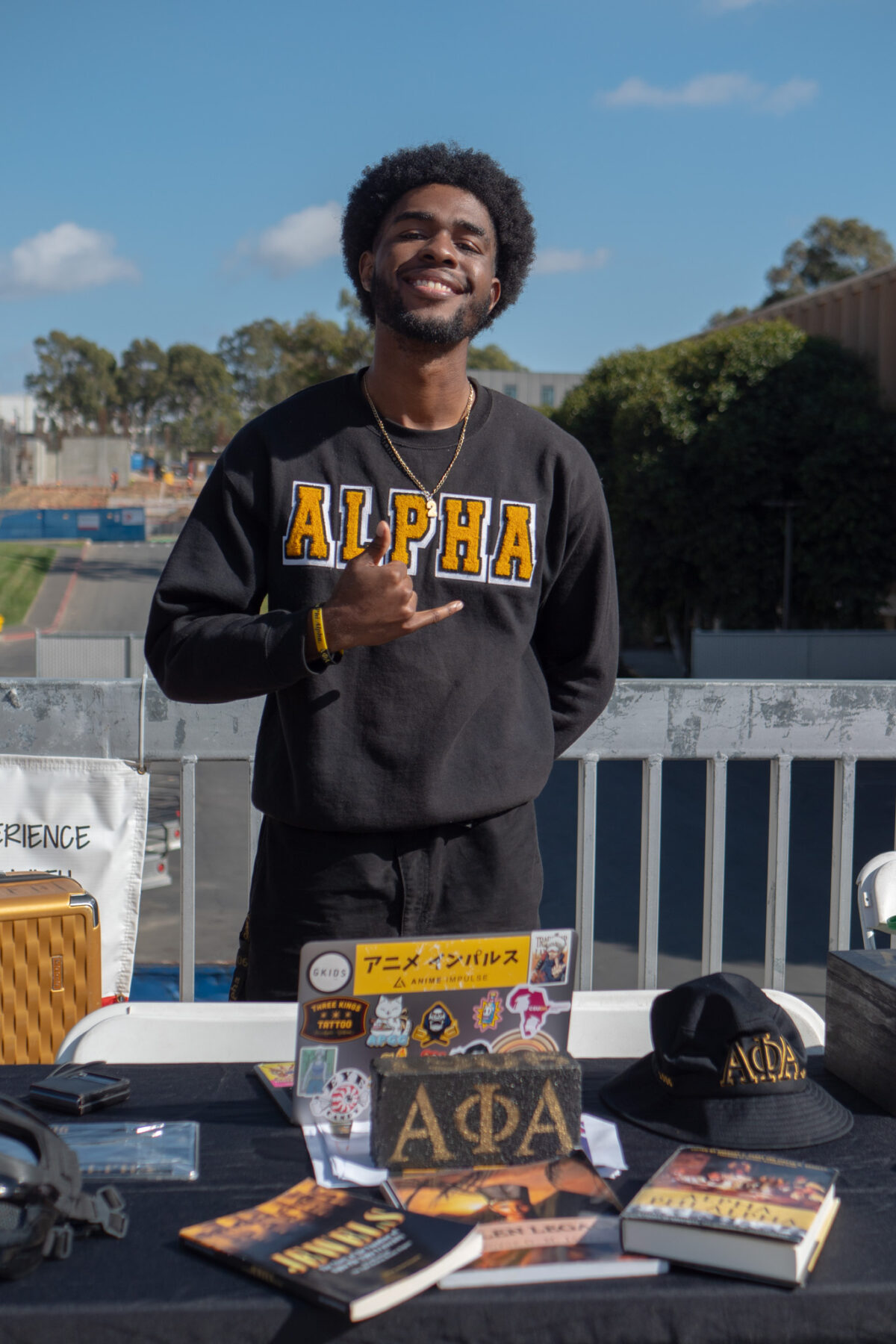 A smiling student wearing an “ALPHA” sweatshirt stands behind a table displaying Alpha Phi Alpha fraternity items, including books, a hat, and a decorated sign, at an outdoor campus event.