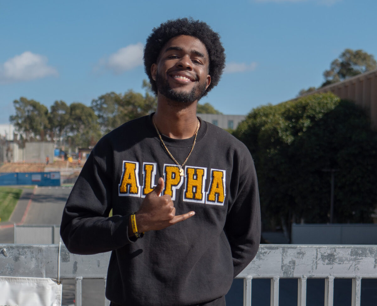 A smiling student wearing an “ALPHA” sweatshirt stands behind a table displaying Alpha Phi Alpha fraternity items, including books, a hat, and a decorated sign, at an outdoor campus event.