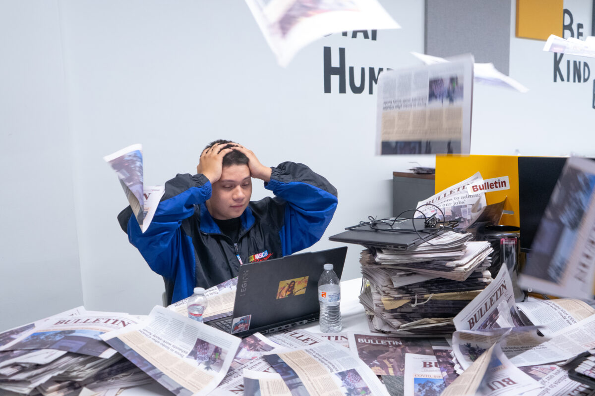 A student sits at a desk with their hands on their head, surrounded by flying newspapers and stacks of paper, looking stressed while working on a laptop in The Bulletin newsroom.