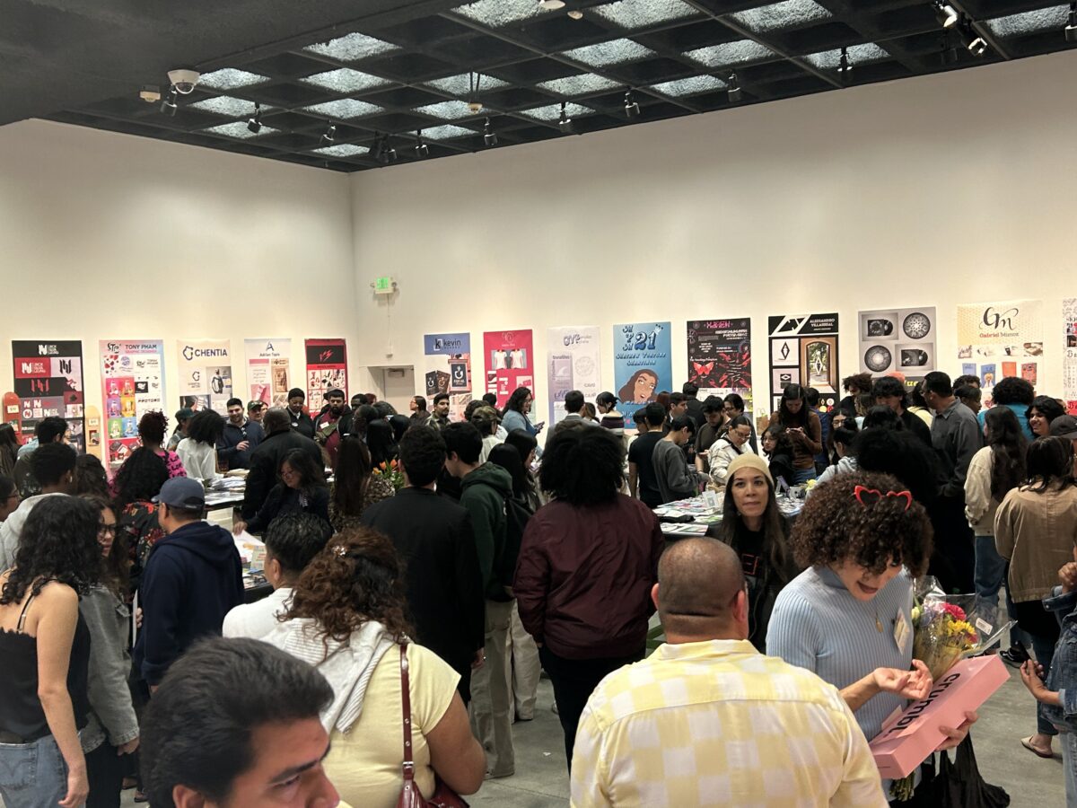 A large crowd gathers inside a gallery space at CSUDH for a student art and design showcase, with posters displayed on the walls and people viewing and discussing work.