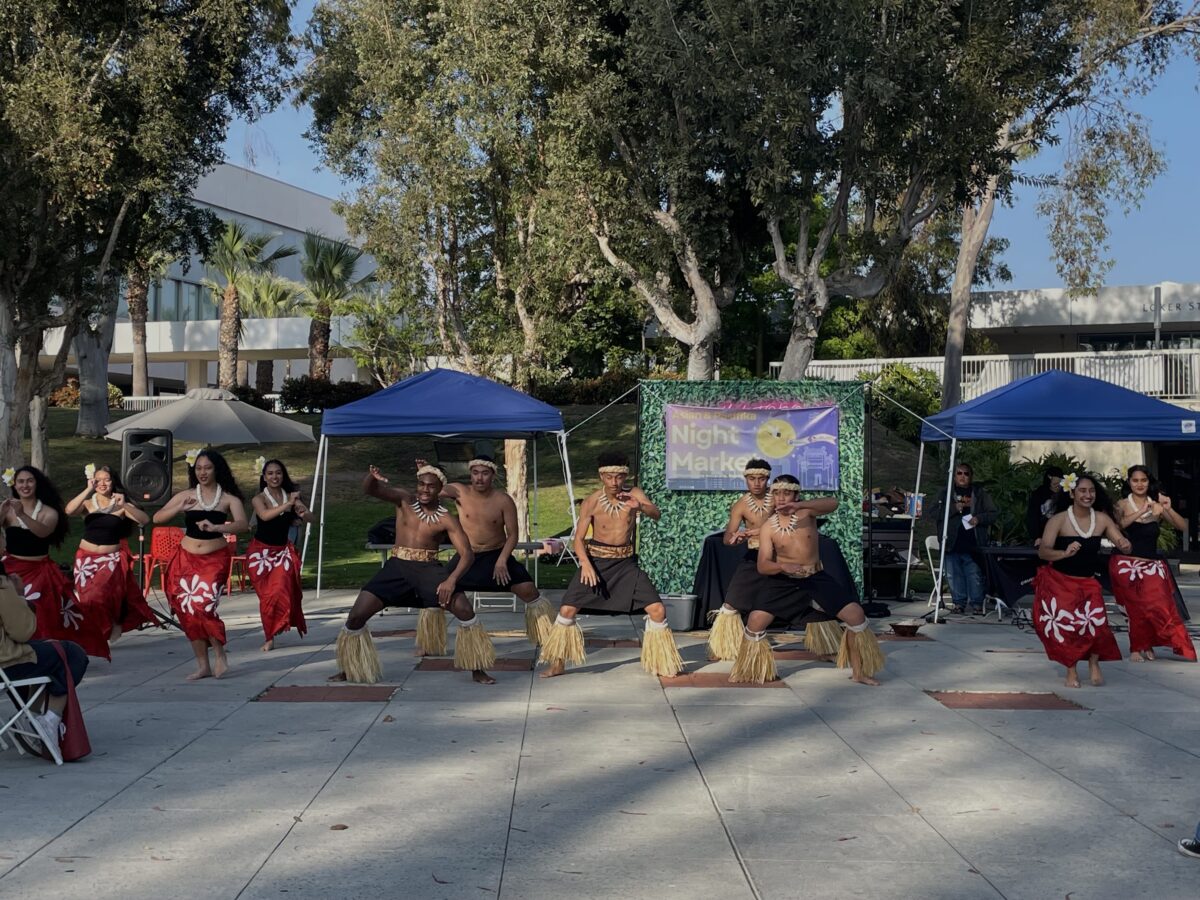 Performers in traditional Polynesian attire dance in front of a “Night Market” banner on the CSUDH campus, with tents and trees in the background.