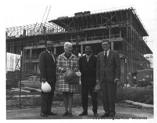 Four people stand in front of a building under construction on the CSUDH campus in a black-and-white archival photo, holding hard hats and posing for the camera.