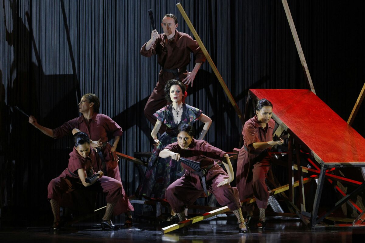 Performers on stage during LA Opera’s production of Ainadamar, with dramatic lighting, red set pieces, and dynamic poses. Dancers in burgundy costumes surround a central figure in a floral dress.