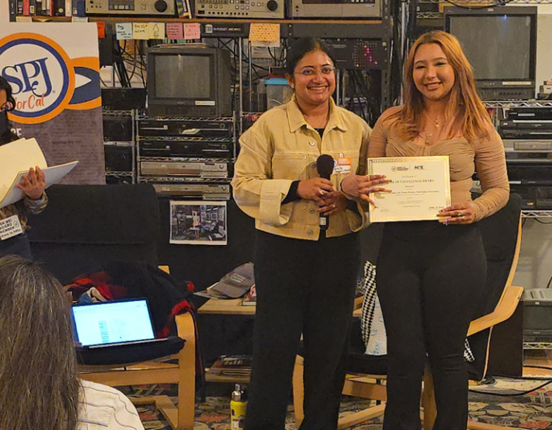 A journalism student holds up their award while standing next to an awards judge in a room filled with CRT televisions and older technologies.